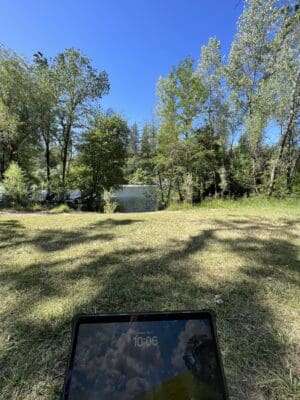 Laptop resting on the grass by the river at Mother Lode River Center, surrounded by trees and a clear blue sky, highlighting the perfect blend of work and nature.