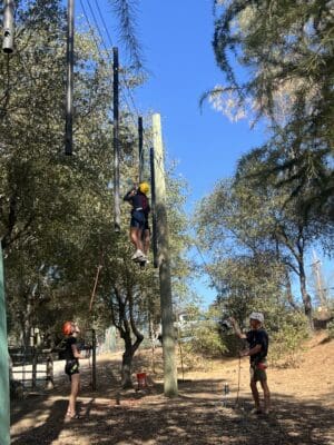 Participants engaging in a ropes course challenge at Mother Lode River Center, with one climber ascending while others provide support and encouragement, embodying teamwork and adventure in the great outdoors.